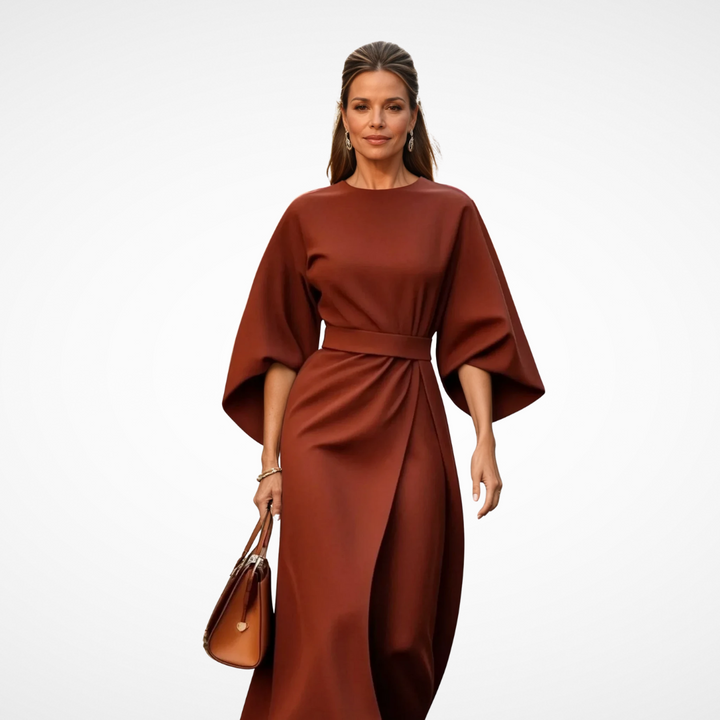 Model dressed in a sophisticated rust midi dress with wide sleeves and waist tie, holding a brown handbag in a studio.