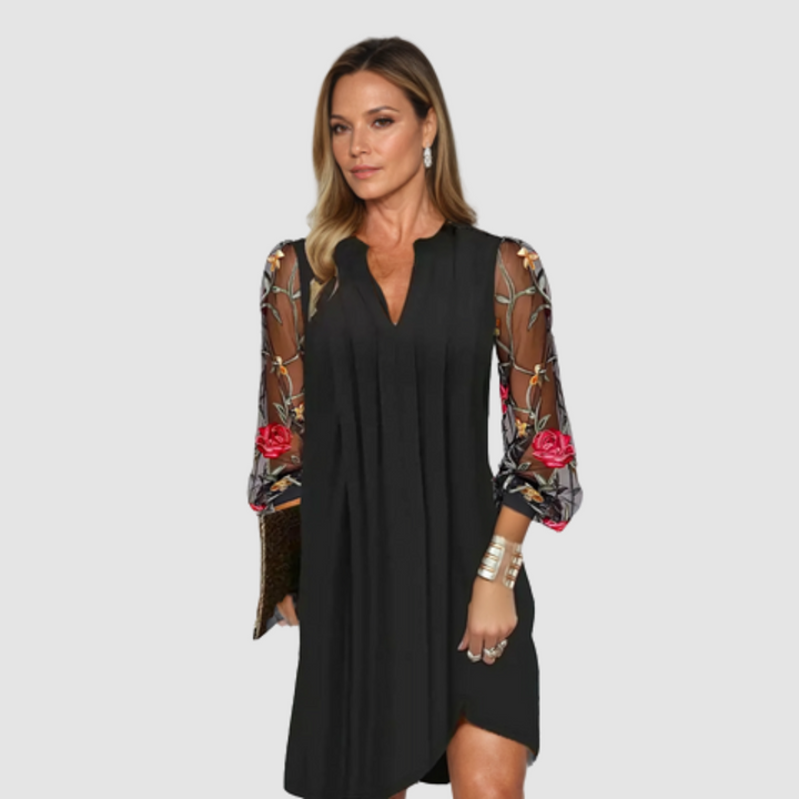 Model wearing a black dress with sheer floral embroidered sleeves, standing against a plain white background.