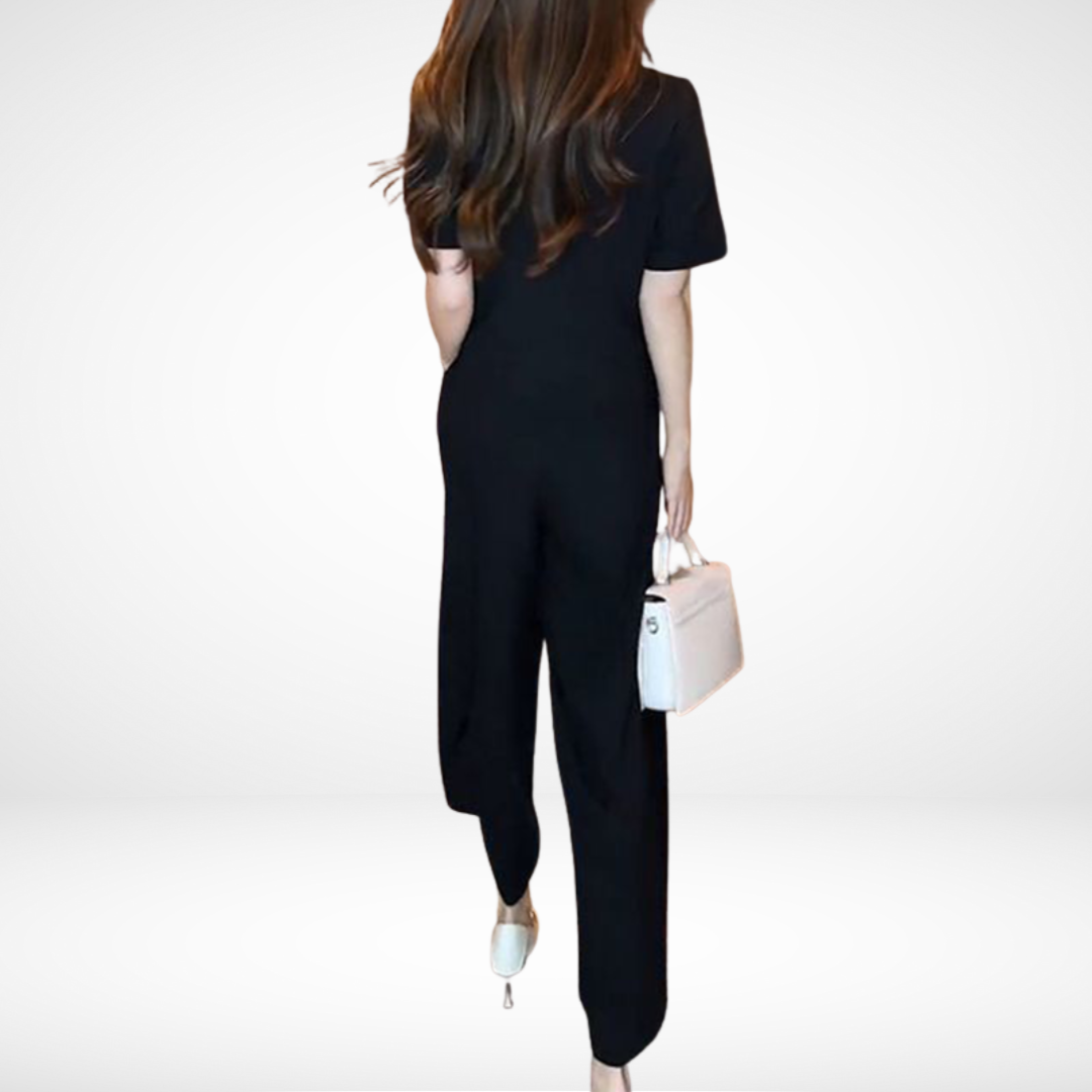 Back view of model in black short-sleeve two-piece set, holding white bag, in white studio setting.