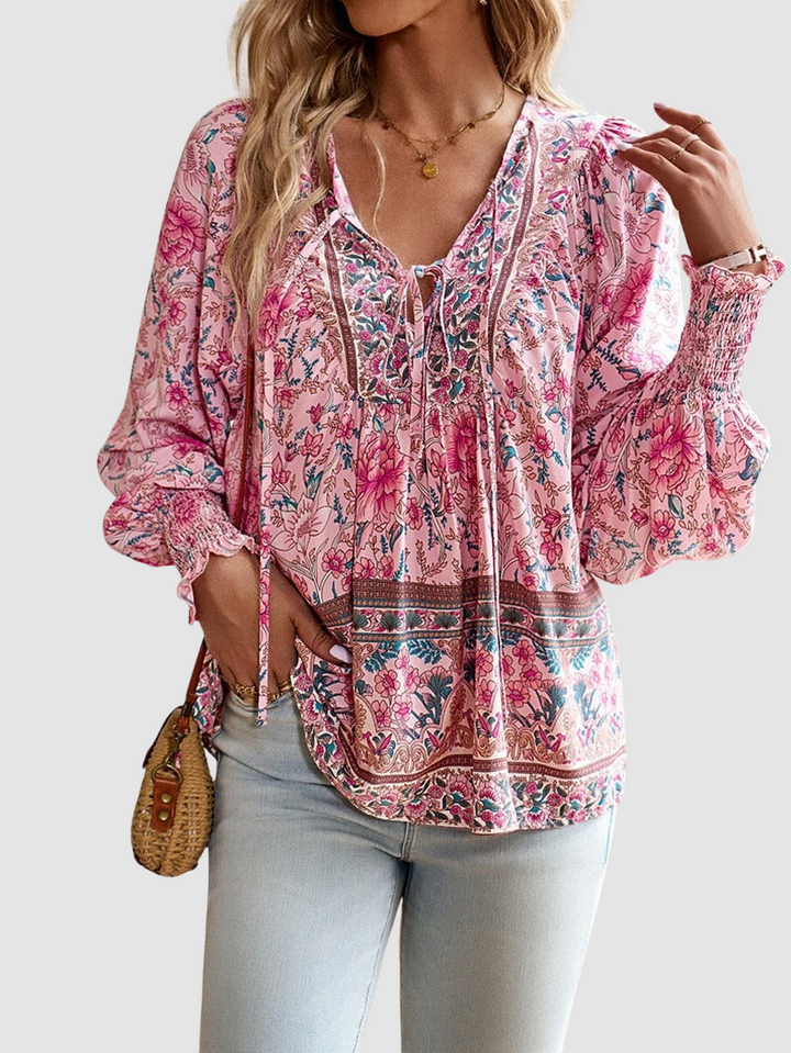 Elegant floral blouse in pink tones with intricate patterns, styled with light denim jeans, against a gray studio backdrop.