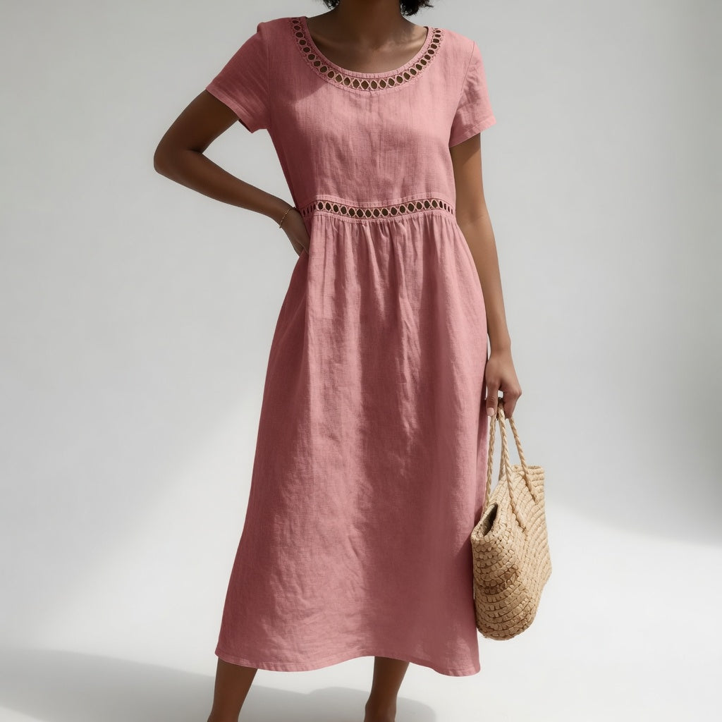 Pink midi dress with eyelet trim and short sleeves, demonstrated by a person with a woven bag. Neutral studio backdrop.