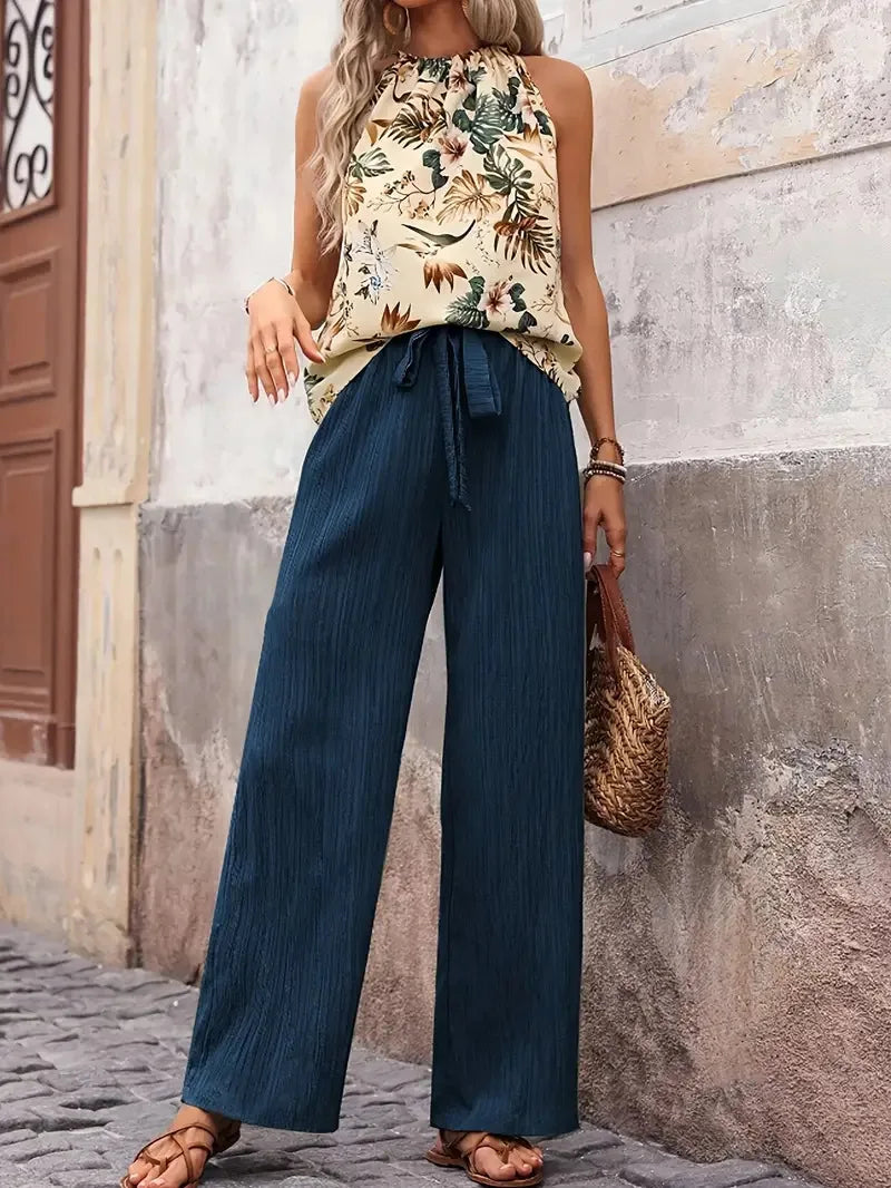 Woman wearing a sleeveless floral top and wide-leg navy pants, holding a woven bag, posed in a rustic street setting.