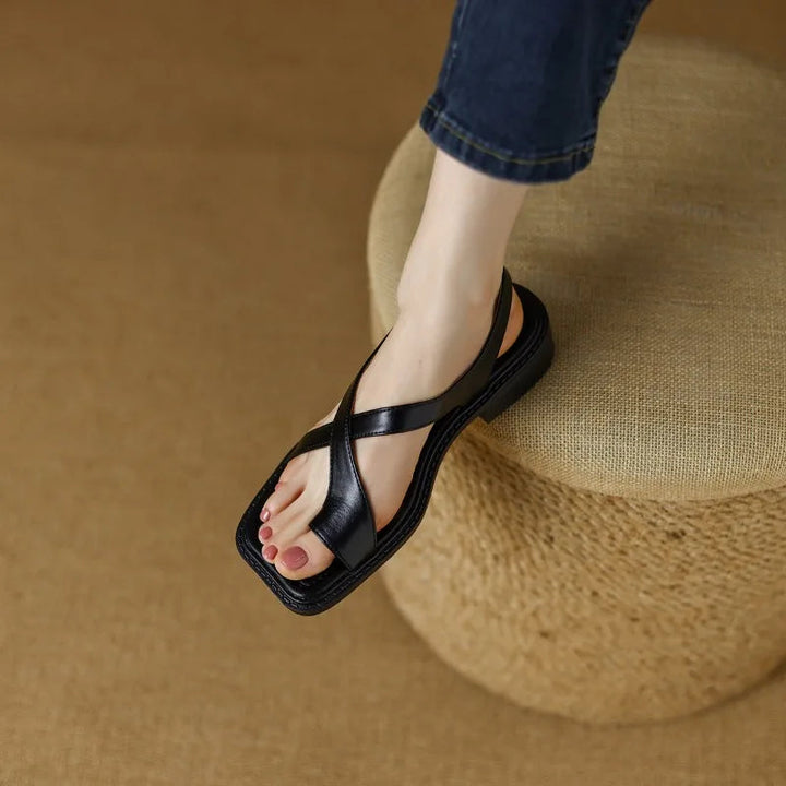 Black leather sandals with crossing straps on a foot, set on a woven ottoman in a warm, indoor environment.