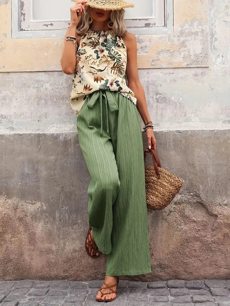 Individual in floral sleeveless top and light green pants, straw hat, and woven bag against a textured urban wall.