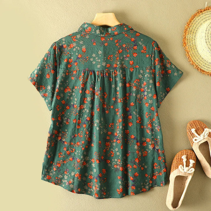Back view of green floral summer shirt on hanger, set against beige wall with straw hat and woven shoes nearby.