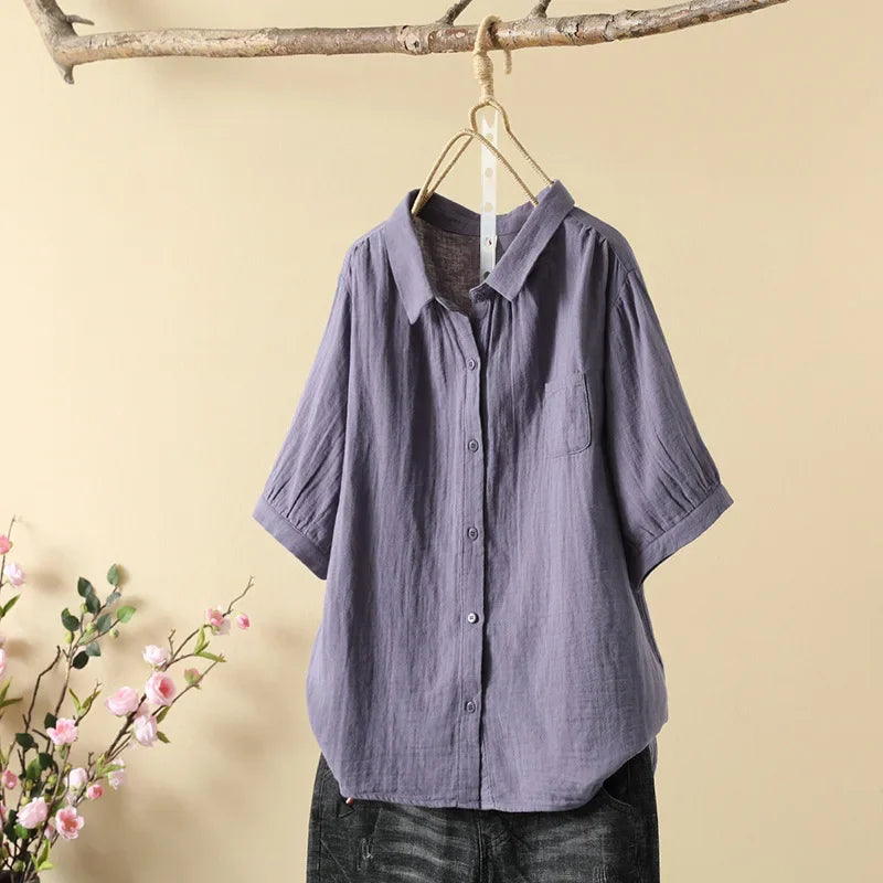Purple casual loose blouse hanging on a wooden branch, with pink flowers nearby, against a light beige background.