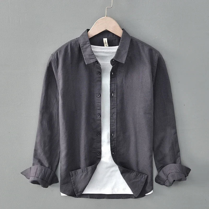 Dark linen shirt with rolled sleeves on hanger, against gray wall.