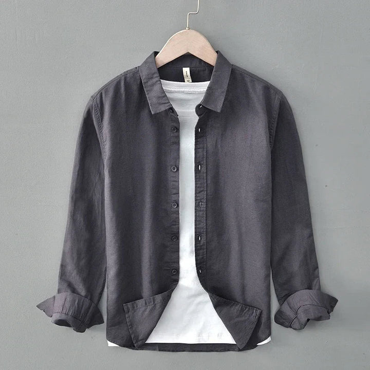 Dark linen shirt with rolled sleeves on hanger, against gray wall.