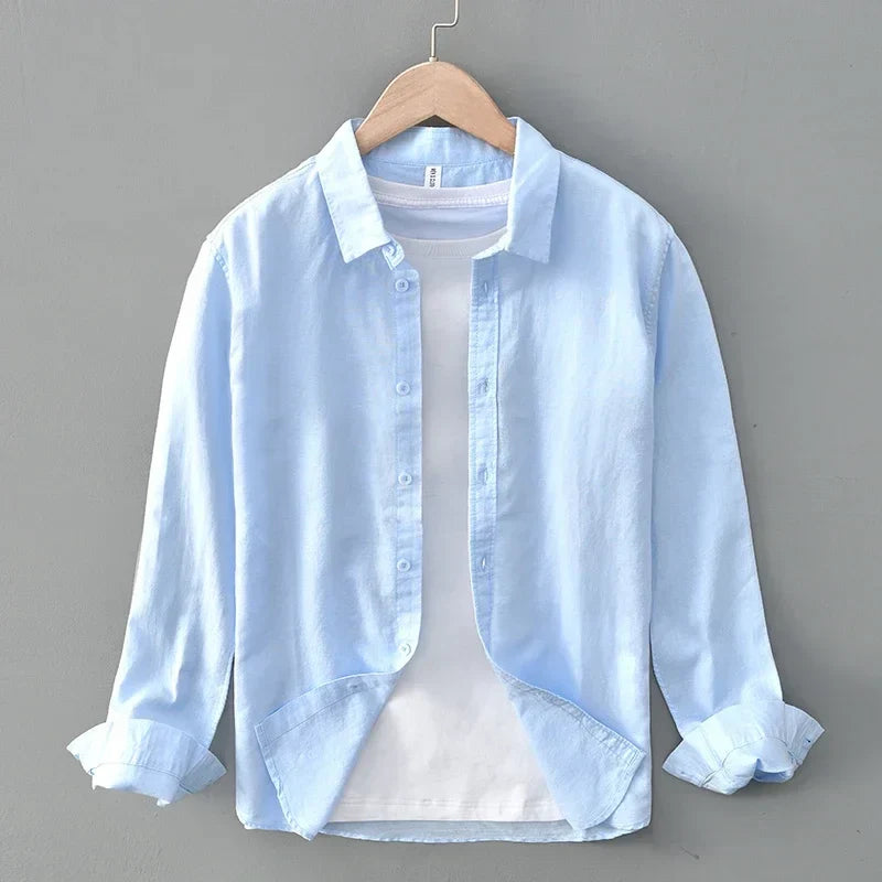 Light blue linen shirt with rolled sleeves on hanger, against gray wall.