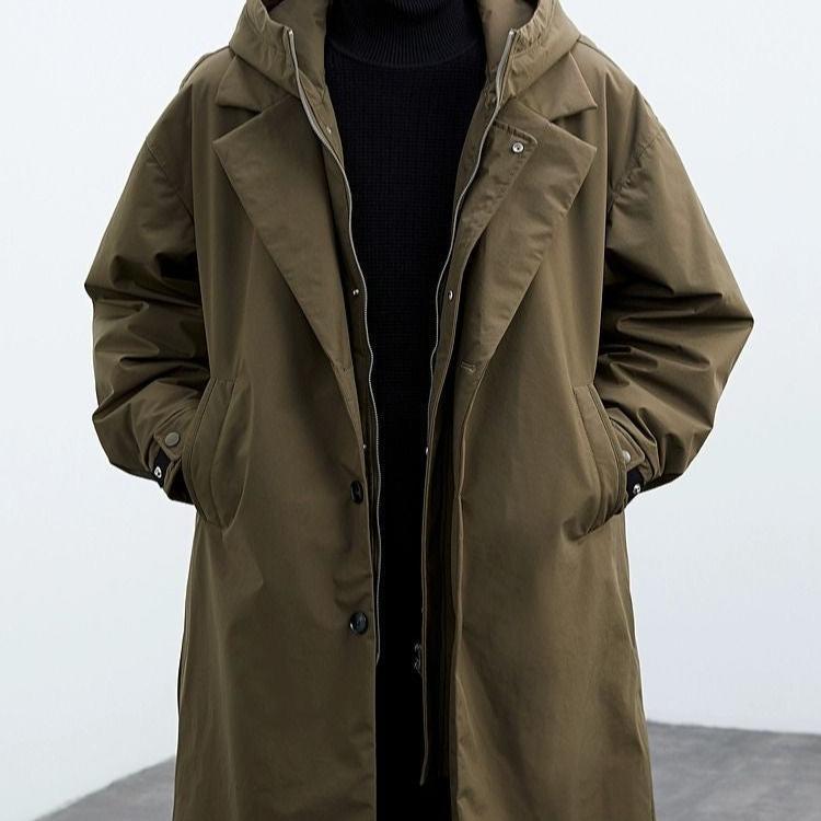 Front view of a brown long coat with hood, hands in pockets, in a gray studio.