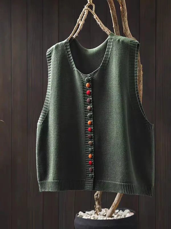 Green sleeveless cardigan with colorful buttons hung on dark wooden background with branch.