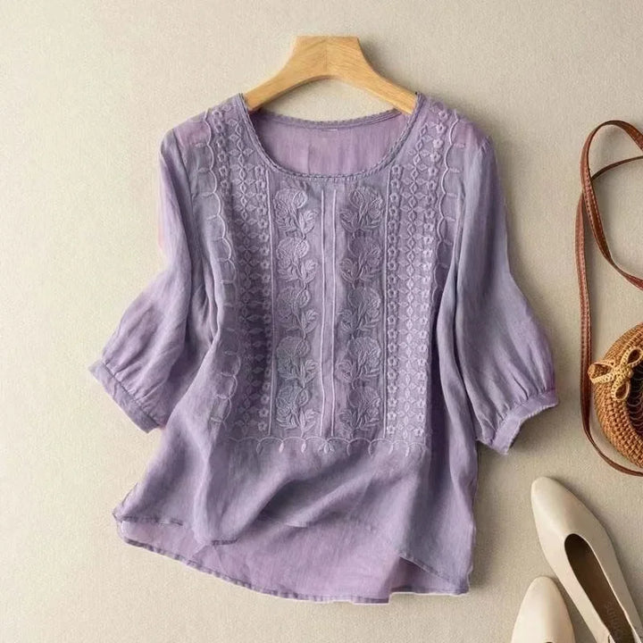 Lavender elegant soft blouse with decorative embroidery on hanger, next to shoes and bag, neutral wall.