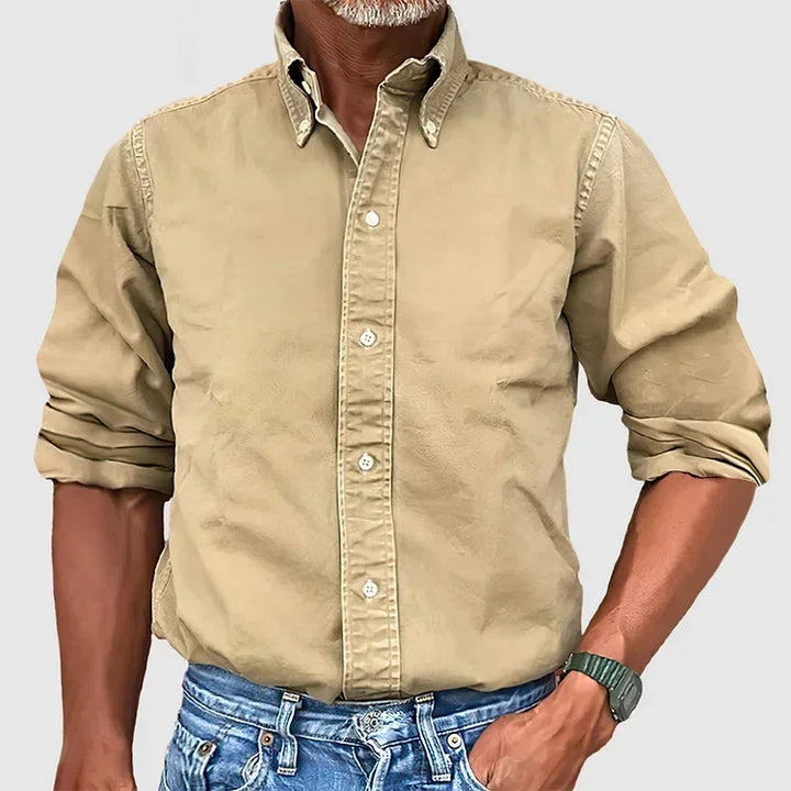Man wearing a beige button-up shirt with rolled sleeves and blue jeans against a plain gray background.