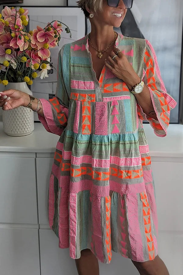 Woman in colorful geometric dress with pink, orange, and green hues. Studio background with flowers and art.