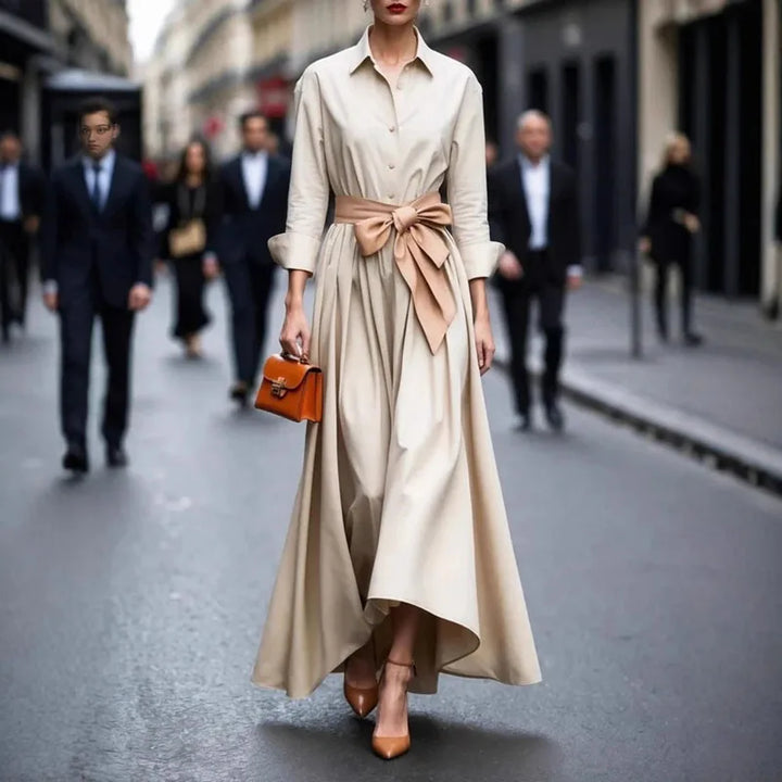 Sophisticated beige maxi dress with a wide bow, complemented by a small orange handbag, walking down a city street.