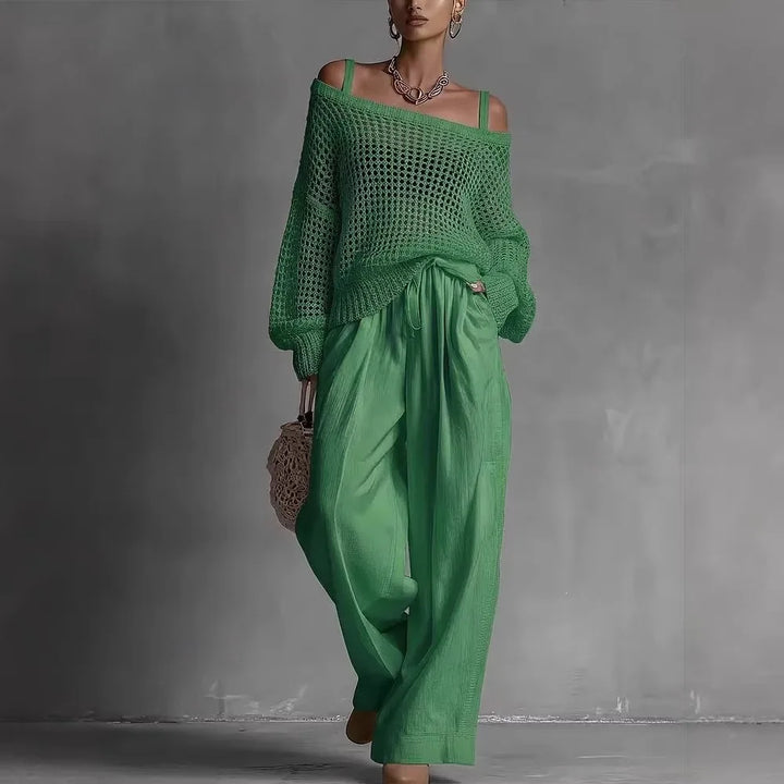Woman in off-shoulder green top and wide green pants, holding woven bag in minimal gray studio.