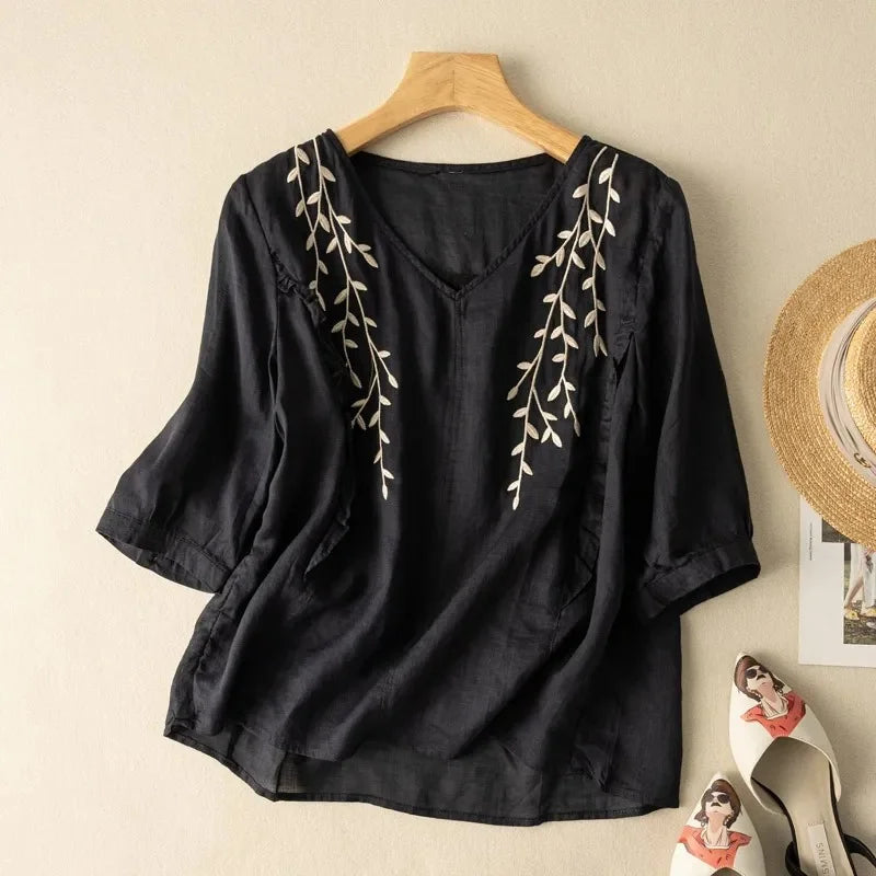 Black loose retro top with white leaf embroidery, displayed on a wooden hanger against a neutral wall background with straw hat and fashion accessories.