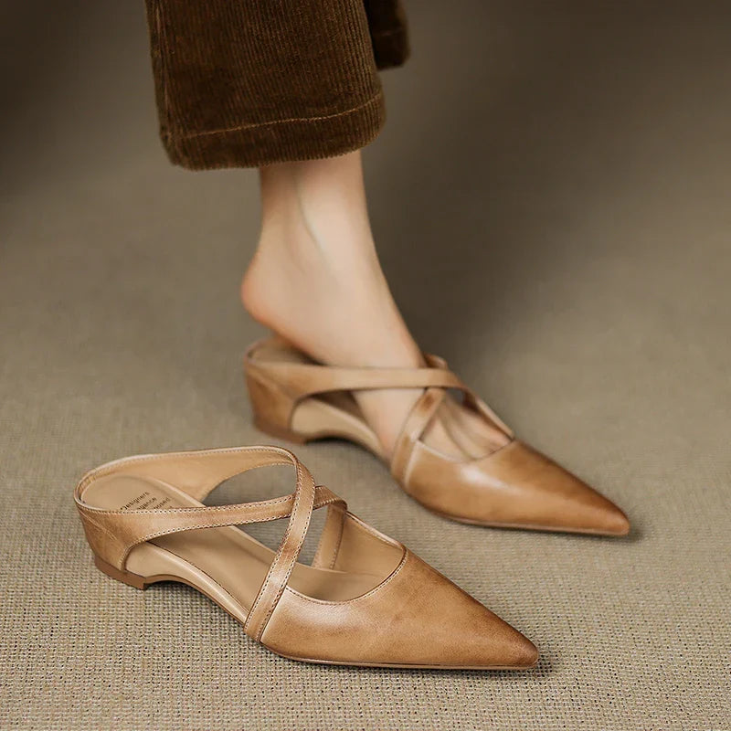 Tan mules with criss-cross straps, one shoe worn and one on the floor.