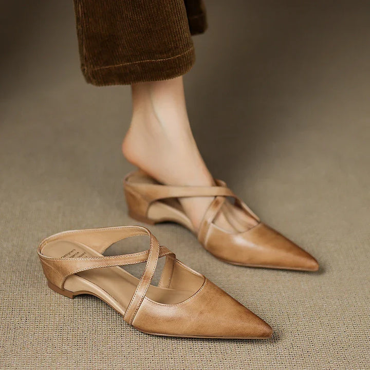 Tan mules with criss-cross straps, one shoe worn and one on the floor.