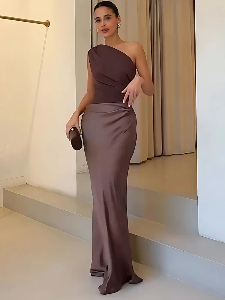Woman in a sleek one-shoulder brown maxi dress, holding a clutch in a minimalist indoor setting with neutral tones.