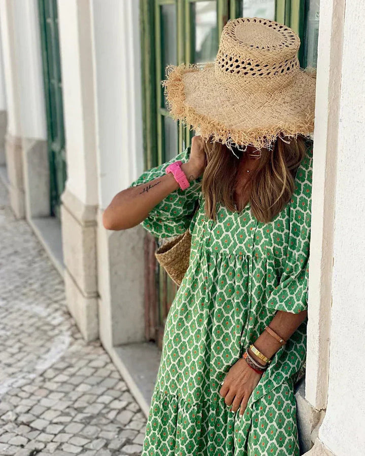 Person in a green bohemian long dress and straw hat leaning against a wall outdoors on a cobblestone street.
