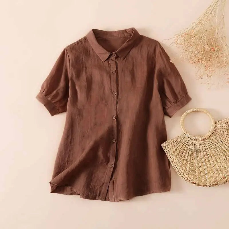 Brown casual button-up shirt with short sleeves, displayed on a neutral background next to a woven handbag and dried flowers.