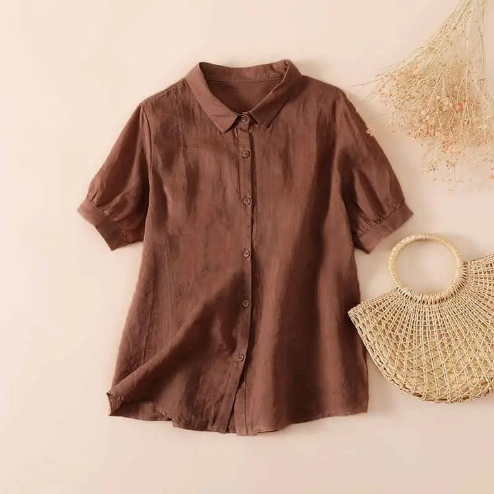 Brown casual button-up shirt with short sleeves, displayed on a neutral background next to a woven handbag and dried flowers.