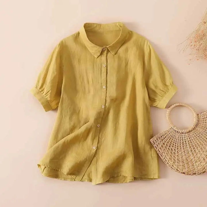 Yellow casual button-up shirt with short sleeves, displayed on a neutral background next to a woven handbag and dried flowers.