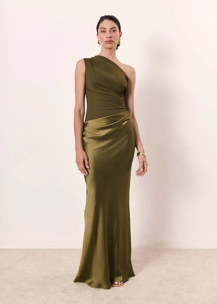 Woman in an olive green one-shoulder maxi dress standing against a plain neutral indoor backdrop.