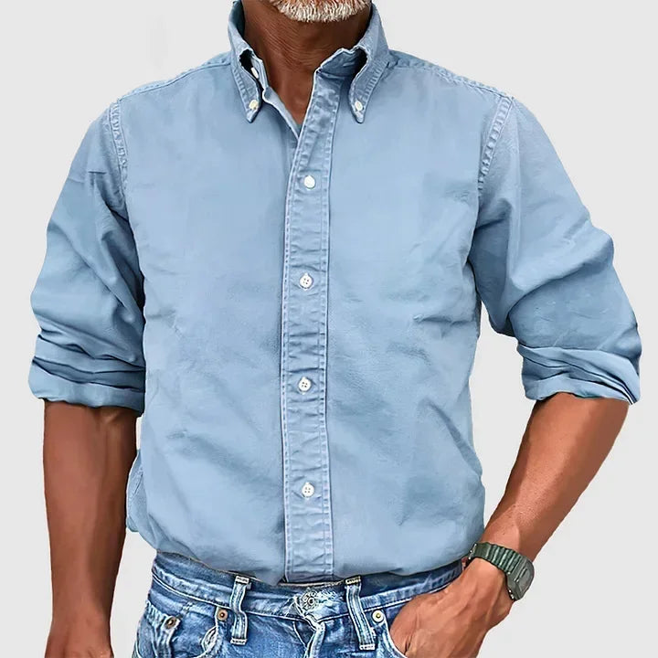 Man wearing a light blue button-up shirt with rolled sleeves and blue jeans against a plain gray background.