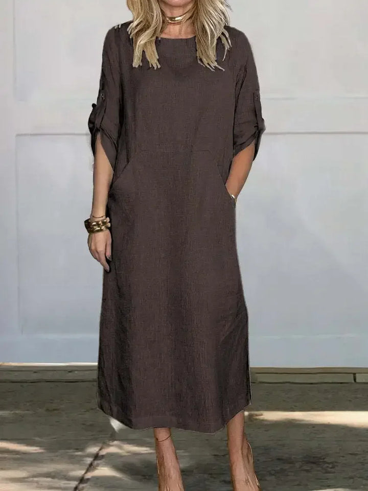 Brown A-line loose fit draped evening dress, studio setting with wooden floor and pale wall background.
