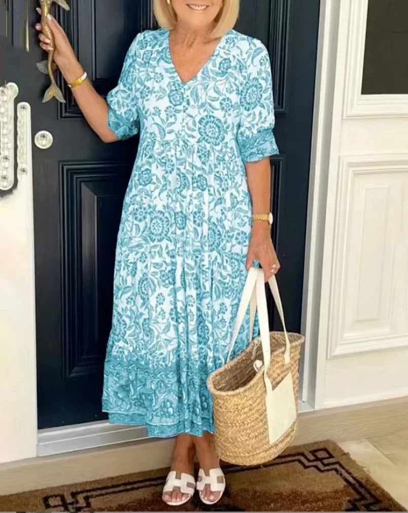 Woman in a turquoise floral V-neck dress with a straw bag, standing by a black door.