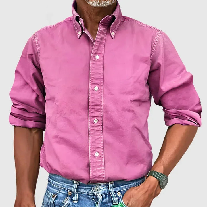 Man wearing a bright pink button-up shirt with rolled sleeves and blue jeans against a plain gray background.