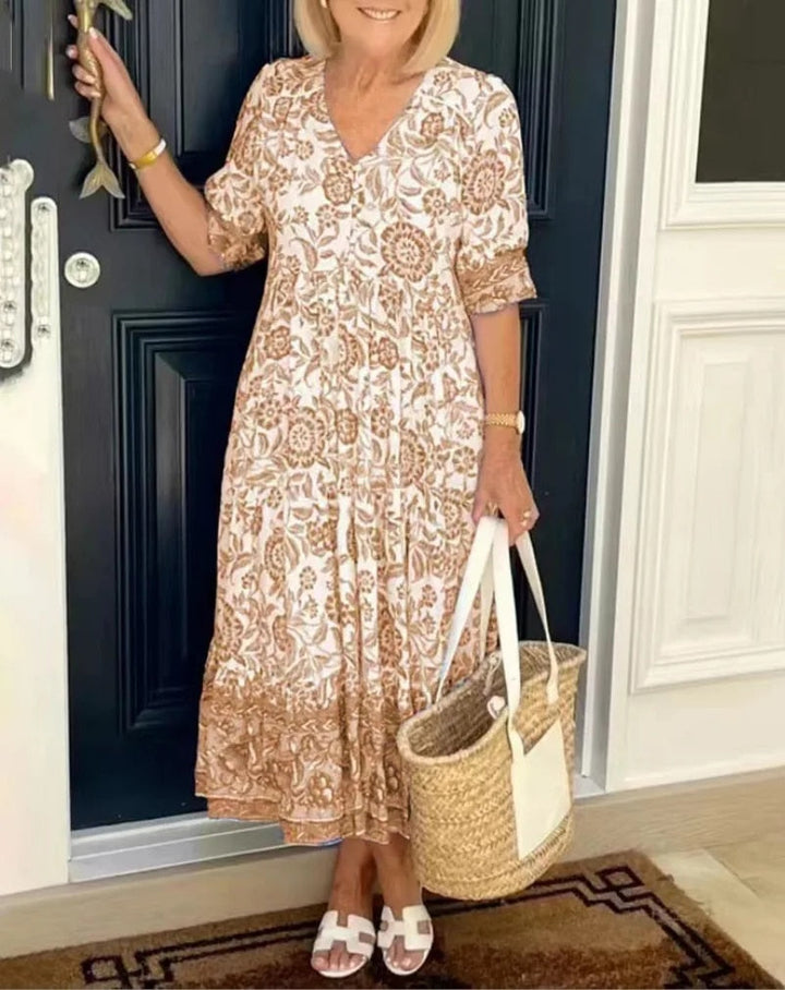 Woman in a brown floral V-neck dress holding a straw tote by a dark door.