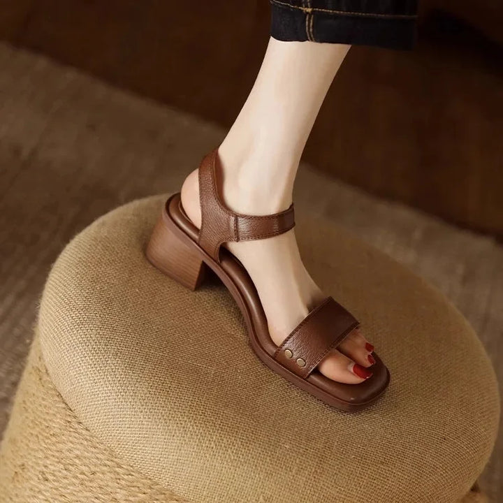 Brown leather sandal with block heel on foot, resting on woven stool, wooden floor background.