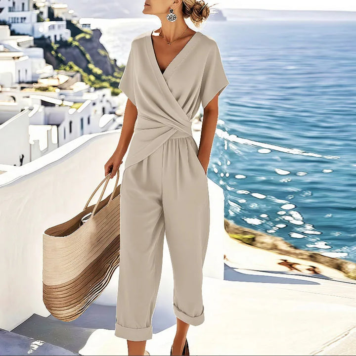Elegant beige two-piece set with wrap top, capri pants, and large woven tote, by seaside with white buildings.