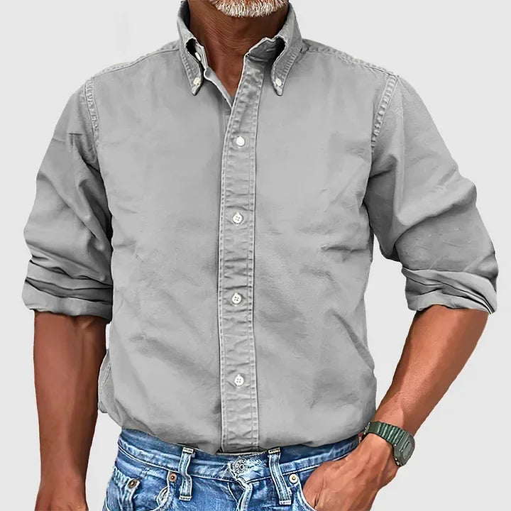 Man wearing a gray button-up shirt with rolled sleeves and blue jeans against a plain gray background.