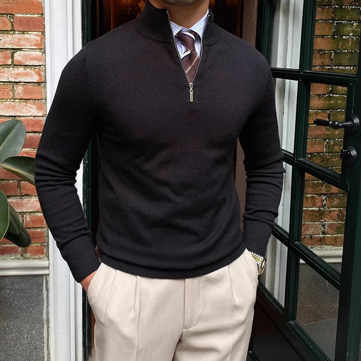 Black half-zip sweater over shirt and tie, worn with beige pants, near a doorway with brick wall background.