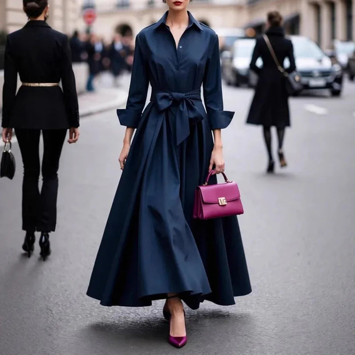 Elegant navy maxi dress with a large bow, paired with a pink handbag and shoes, on a city street.