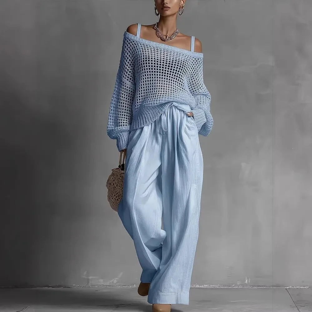 Woman in off-shoulder blue top and wide blue pants, holding woven bag in minimal gray studio.