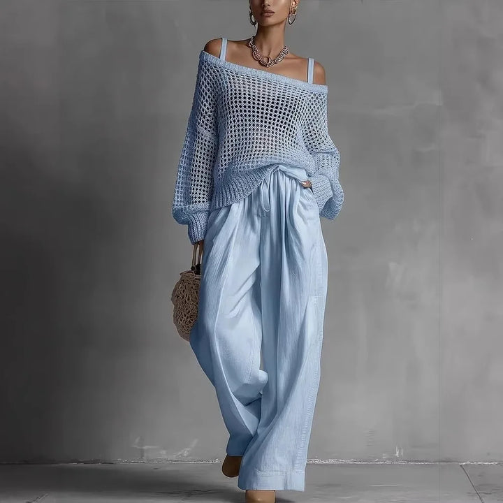 Woman in a blue, off-shoulder knit top and matching flowing trousers, holding a netted bag, in a gray studio setting.