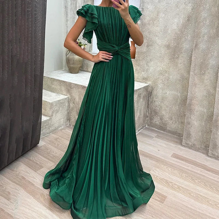Elegant green pleated evening dress with ruffled sleeves, modeled in a chic indoor setting with modern decor.