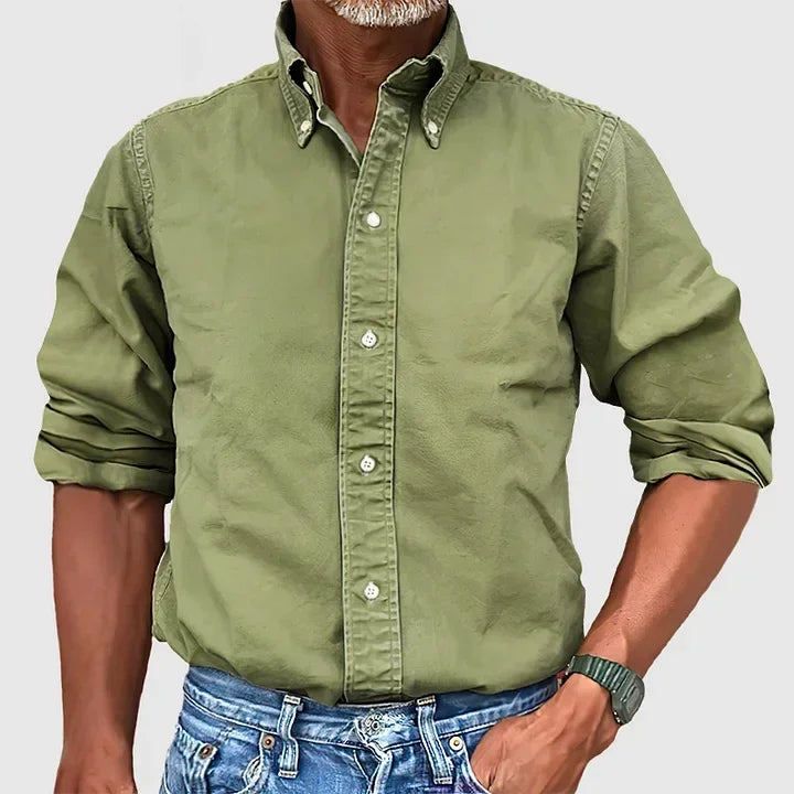 Man wearing a green button-up shirt with rolled sleeves and blue jeans against a plain gray background.