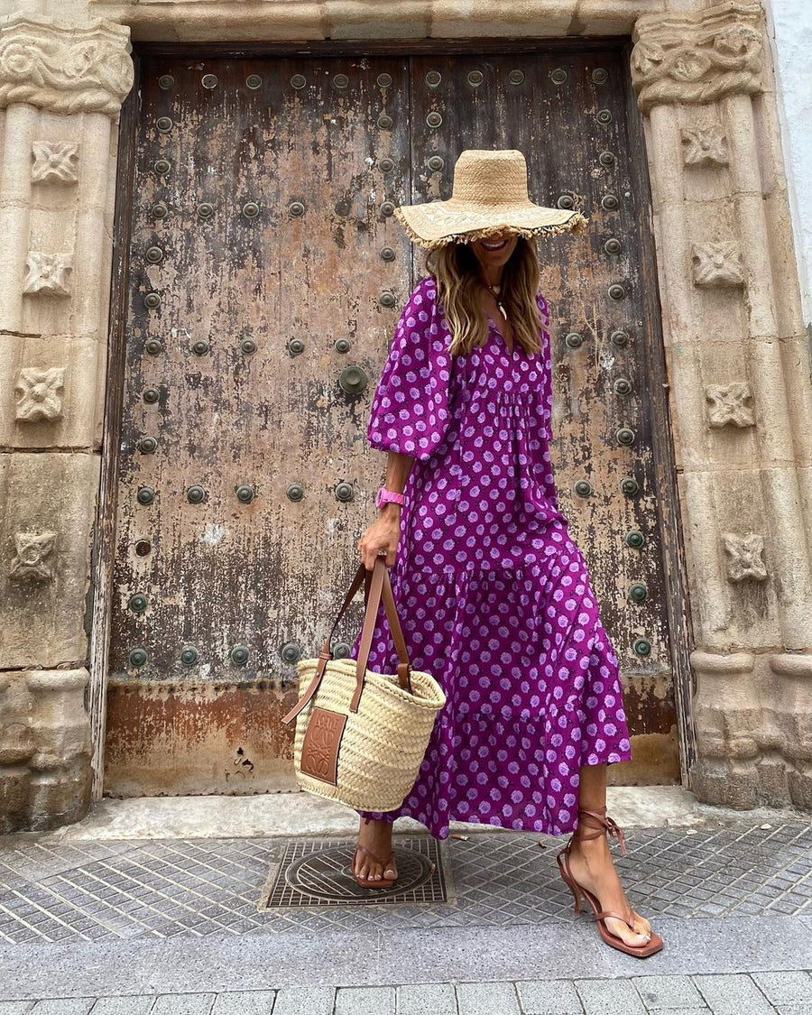 Purple patterned bohemian long dress and straw hat in front of ornate wooden doors outdoors.