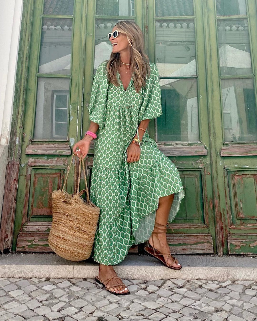 Green bohemian long dress with geometric pattern, worn by a person holding a straw bag, standing in front of rustic green doors.