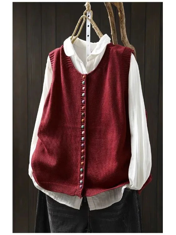 Red sleeveless cardigan with colorful buttons over white blouse hung on dark wooden background.