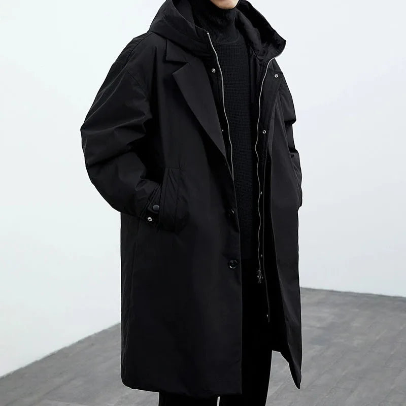 Person wearing a black long coat with hood, standing in a gray studio setting.