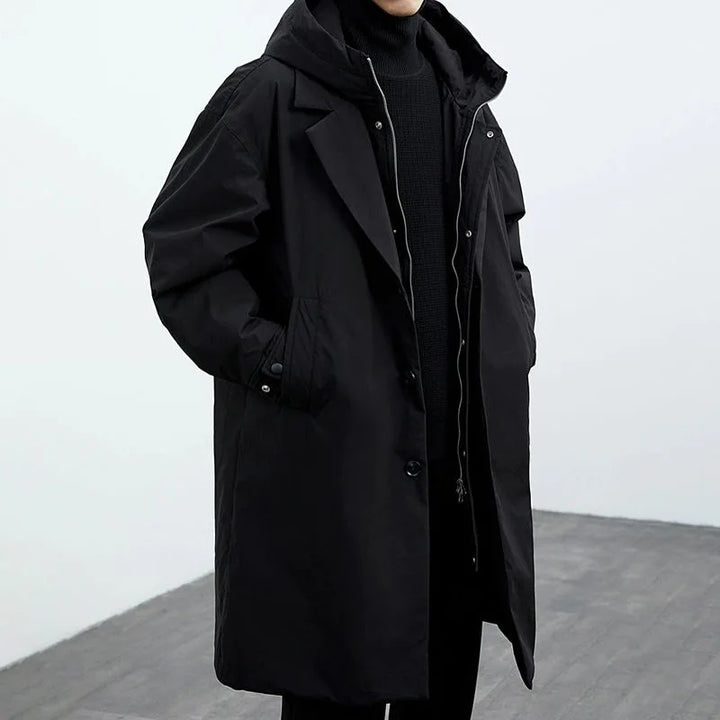 Person wearing a black long coat with hood, standing in a gray studio setting.