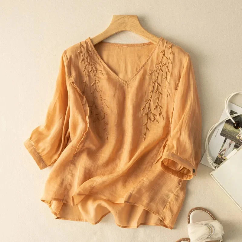 Peach loose retro top with subtle leaf embroidery, hung on a wooden hanger against a neutral wall, with sandals and bag nearby.