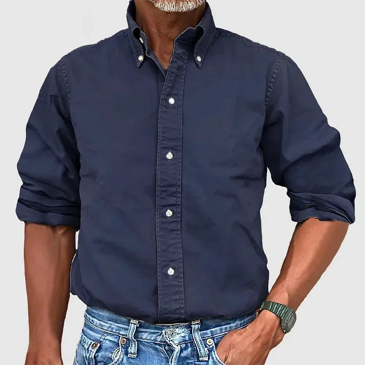 Man wearing a navy blue button-up shirt with rolled sleeves and blue jeans against a plain gray background.